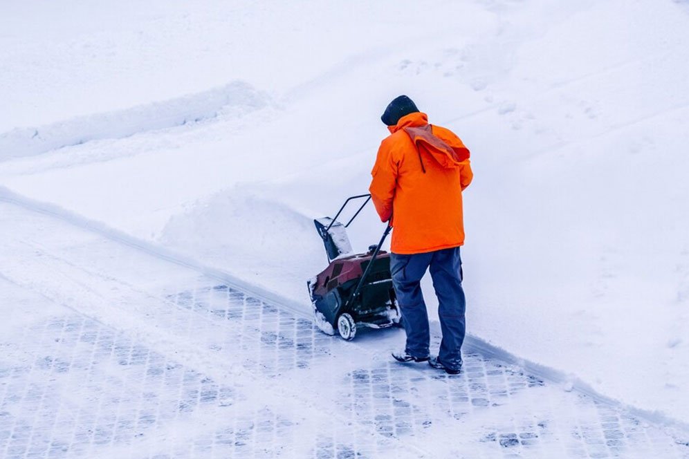A man operates a snow blower, clearing a path through freshly fallen snow in a residential area.
